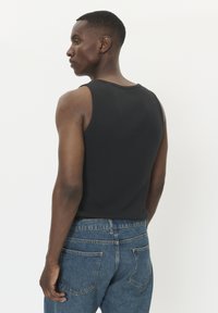 Uomo che indossa una canottiera nera senza maniche e jeans blu, voltato di lato di fronte a uno sfondo bianco semplice.