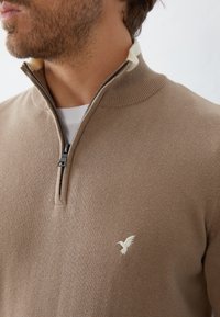 Beige stickad tröja med dragkedja i kragen, med en diskret broderad fågel-logotyp på bröstet och en texturerad tygfinish.