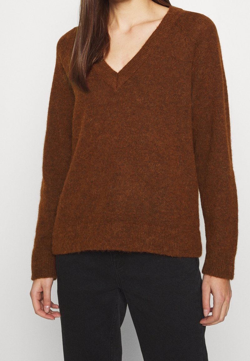 Pull brun à col en V fabriqué en matière douce et texturée avec des manches longues. Présente une coupe décontractée et un ourlet côtelé, assorti à un jean noir.