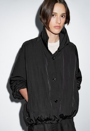 Un mannequin avec des cheveux bruns courts porte une chemise veste noire oversize, se tenant contre un fond gris clair, mains dans les poches.