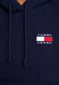 Felpa con cappuccio in pile blu navy con superficie testurizzata, caratterizzata da un logo quadrato con "TOMMY JEANS" e blocchi di colore rosso, bianco e blu.