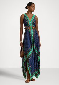 Γυναίκα που φοράει μπλε μακρύ φόρεμα χωρίς μανίκια με πράσινο και καφέ γεωμετρικό σχέδιο, καφέ ζώνη, σανδάλια και κρατά μια μικρή καφέ τσάντα.