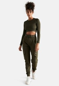 Femme aux cheveux bouclés porte un crop top côtelé vert olive et un pantalon cargo assorti avec ceinture et baskets blanches, debout sur fond blanc.