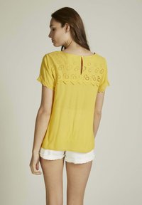 Blouse jaune avec des manches courtes en dentelle festonnée, présentant un dos découpé décoratif et un tissu doux et léger. Associée à un short blanc.