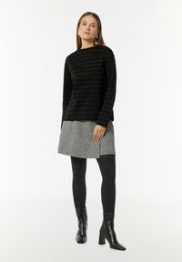Czarny sweter w wzór houndstooth, szara spódnica o fakturowanym wykończeniu z ozdobami, czarne rajstopy i botki za kostkę. Modelka pozuje na neutralnym tle.