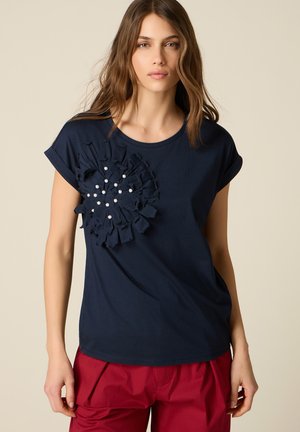 Giovane donna con capelli lunghi indossa un top blu navy a maniche corte con fiore texturizzato con perline bianche, abbinato a pantaloni rossi, sfondo neutro.