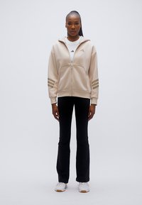 Μπεζ φούτερ με φερμουάρ και τρεις οριζόντιες ρίγες στα μανίκια, συνδυασμένο με μαύρο παντελόνι και λευκά αθλητικά παπούτσια, με casual εφαρμογή και λεία υφή.