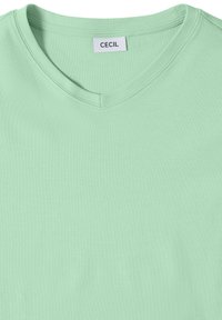 Chemise vert clair à col en V avec une étiquette blanche portant l'inscription "CECIL" cousue à l'intérieur du col.