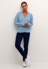 Hellblauer gestrickter Cardigan mit V-Ausschnitt und Knopfverschluss, der ein strukturiertes Muster aufweist. Tragen über dunklen Jeans und weißen Sneakers.