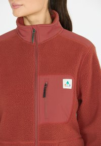 Rote Fleecejacke mit hohem Kragen, Reißverschluss und einer Brusttasche. Mit einem kontrastierenden glatten Einsatz und Logo. Strukturierten Stoff.