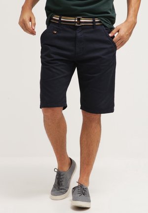 Homme portant un short chino bleu marine avec une ceinture rayée, une chemise vert foncé et des chaussures décontractées grises à lacets, debout sur un fond blanc.