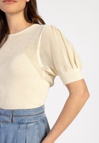 Krémový top s texturovaným síťovým designem a nadýchanými krátkými rukávy, doplněný o žebrované detaily, v kombinaci se světle modrou džínsovou sukní.