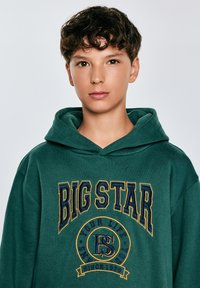 Felpa verde in cotone con un grande logo "BIG STAR" in giallo e blu navy. Dotata di cappuccio con cordini e vestibilità casual.
