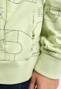 Sudadera verde claro con patrones geométricos negros, puños acanalados y una pequeña etiqueta de logo amarilla en el bolsillo. Textura suave y corte casual.