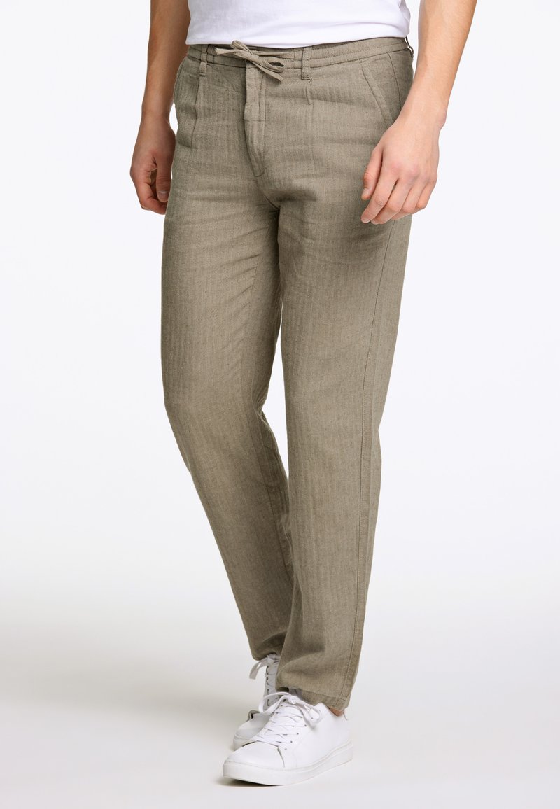 Helle beige, gewebte Stoffhose mit lockerer Passform, Kordelzugbund und seitlichen Taschen. Kombiniert mit weißen Sportschuhen.