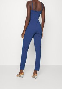 Mörkblå jumpsuit med spaghetti-stöd, figursydd midja, raka ben och bakzip, som matchas med metalliska klacksandaler.