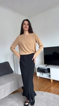 Γυναίκα που φοράει μπεζ μακρυμάνικη μπλούζα και μαύρη ριμπ μάξι φούστα, στέκεται σε μοντέρνο σαλόνι με τηλεόραση και καναπέ.