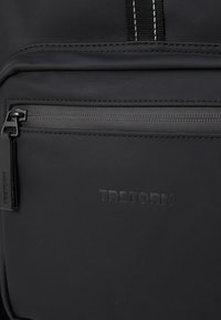 Poche avant du sac à dos Tretorn noir avec fermeture éclair et nom de marque en relief, gros plan montrant les détails de couture et la texture du tissu.