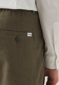 Olive green trousers with a textured fabric, featuring a single button closure, pocket with a sewn label, paired with a white shirt.