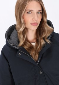 Donkerblauwe jas met een grijze fleece-voering in de capuchon, uitgerust met een ritssluiting en drukknoopsluiting. Glad oppervlak met een strak ontwerp zonder zichtbare patronen.