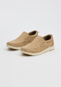 Zapatos de ante beige sin cordones con paneles laterales elásticos, que cuentan con una suela de goma blanca y costuras decorativas a lo largo del borde superior.