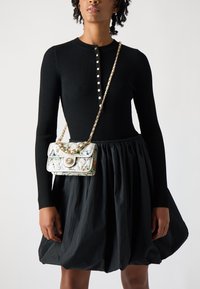 Haut noir à côtes à manches longues avec fermeture à boutons, associé à une jupe noire bouffante. Petit sac blanc avec un motif floral et une chaîne dorée.