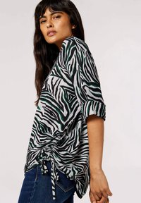 Grijs top met zwarte en groene zebra strepen, korte mouwen en een trekkoord detail in de taille. Draagt met blauwe jeans.