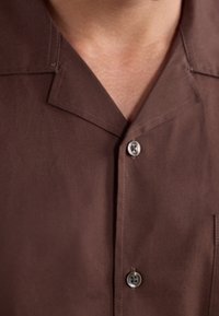 Brown short-sleeved shirt with a notched collar, featuring two buttons and a smooth fabric texture. No visible patterns or accents.