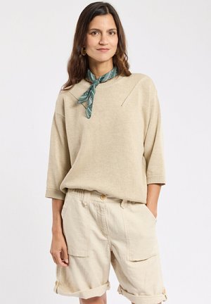 Femme portant un pull en maille beige, un short cargo crème aux ourlets retroussés et un foulard vert à motifs autour du cou, debout avec une main dans la poche.