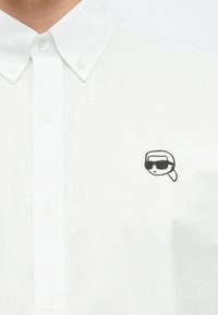 Biała koszula na guziki wykonana z bawełny. Na lewej stronie klatki piersiowej znajduje się czarny haft przedstawiający postać w okularach przeciwsłonecznych.