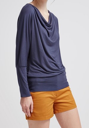 Persoon draagt een donkerblauw shirt met lange mouwen en een gedrapeerde halslijn, gecombineerd met mosterdgele korte broek, tegen een effen lichte achtergrond.