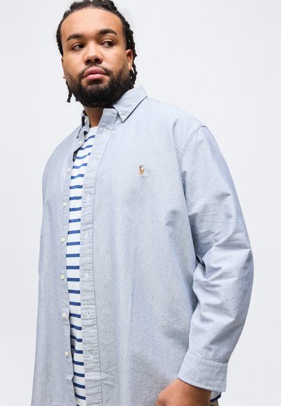 Άνδρας με πλεξούδες που φοράει ένα ανοιχτό μπλε πουκάμισο πάνω από μια μπλε και λευκή ριγέ κοντομάνικη μπλούζα, κοιτάζει προς το πλάι μπροστά σε απλό φόντο.