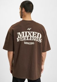 Brun bomull t-skjorte med fet hvit tekst "MIXED FEELINGS" på baksiden, med avslappet passform og kortermede ermer.