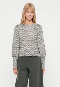 Νεαρή γυναίκα με ξανθά μαλλιά, ντυμένη με μια υφασμάτινη γκρίζα μπλούζα με μακριά μανίκια και ψηλόμεσες φαρδιές γκρίζες παντελόνες, που στέκεται μπροστά σε λευκό φόντο.