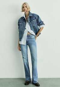 Denim-takki ja vaaleansiniset farkut, joissa on rento istuvuus, kuluneet helmat ja valkoinen nappipaita alla. Tummat kengät viimeistelevät asun.