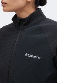 Γυναίκα που φοράει μαύρο μπουφάν Columbia με φερμουάρ, ψηλό γιακά και λογότυπο στο στήθος, με τα μαλλιά της πιασμένα σε κότσο.