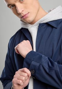 Navy blue jacket with a button cuff, layered over a gray hoodie. Smooth fabric with a structured design and visible stitching details.