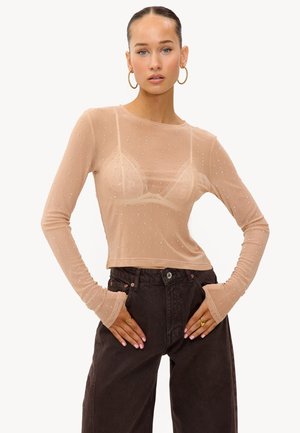 Femme avec les cheveux plaqués en arrière portant un haut long à manches transparentes couleur chair orné de rhinestones, un soutien-gorge en dentelle, des boucles d'oreilles en anneau doré, et un jean noir taille haute.