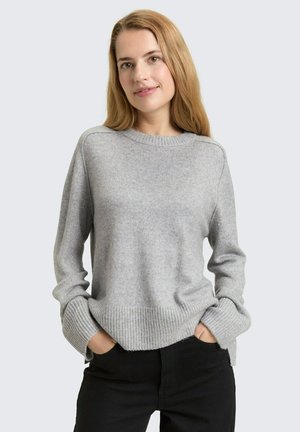 Frau mit langen blonden Haaren, die einen hellgrauen gestrickten Pullover und schwarze Hose trägt, mit den Händen in den Taschen, stehend vor einem einfarbigen Hintergrund.