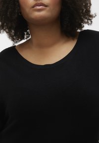 Pull en tricot noir avec un large col rond, présentant une surface texturée et une coupe décontractée. Gros plan montrant les détails du tissu.