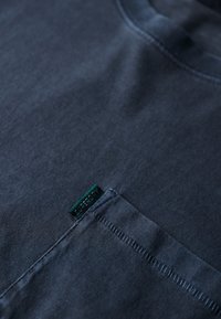 Granatowa koszulka, gładka w dotyku, okrągły dekolt, z małą zieloną metką z napisem "Superdry" w pobliżu dołu oraz widocznymi detalami szycia.