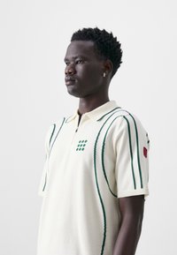Cream short-sleeve polo shirt with green stripes and polka dot pattern on the chest. Features a zip collar and red logo patch on sleeve.