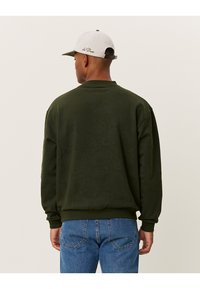 Felpa verde con un motivo testurizzato, collo a giro, e spalle scese, abbinata a jeans blu e un cappellino grigio chiaro.