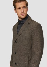 Brauner Textur-Mantel mit einem eingeklappten Revers, drei schwarzen Knöpfen und zwei Seitentaschen, über einer dunklen, mit Reißverschluss versehenen Innensjacke.