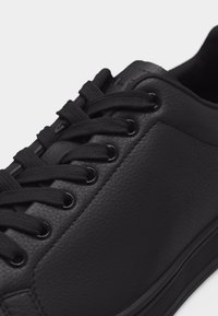 Baskets en cuir noir avec surface texturée, lacets noirs passés à travers six œillets en métal, et semelle en caoutchouc sur fond blanc.