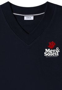 T-shirt bleu marine col V avec texte blanc "Mer & Soleil", graphisme corail rouge et slogan "Sea Sand Sunshine" sur la poitrine, étiqueté "CECIL" à l'intérieur du col.