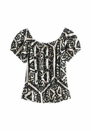 Femme portant une robe longue noire et blanche à motif, épaules nues, debout près d'un balcon blanc avec l'océan en arrière-plan.