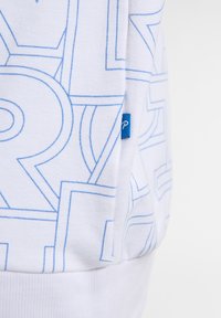 Witte sweatshirt met een blauw geometrisch patroon, zachte textuur, geribbelde manchetten en een klein blauw label aan de zijkant.
