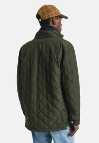 Gepolsterte olivgrüne Jacke mit hohem Kragen und gefüttertem Innenbereich, ausgestattet mit einem Knopfverschluss und einer hellbraunen Schirmmütze im Karomuster.