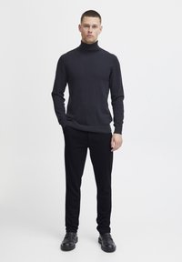 Dunkler Rollkragenpullover aus glattem Strickstoff, kombiniert mit schwarzen, maßgeschneiderten Hosen und schwarzen Schuhen. Einfaches, monochromatisches Design.